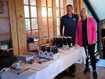 Two people stand behind a table with homemade products, including jars and bottles, in a warmly lit indoor setting with wooden walls and windows.
