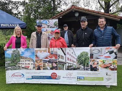 Five people stand outdoors holding a banner with images of businesses. Trees and a small building are in the background, suggesting a casual event.