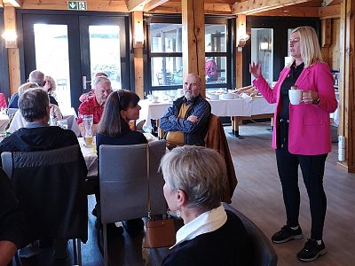 Several people sitting at a table in a rustic room, a woman in a pink jacket is standing and speaking to the group while holding a white mug.