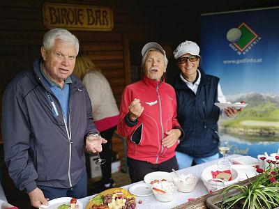 Three seniors outdoors, enjoying food and beverages at a table. They are casually dressed, smiling, with mountains visible in a backdrop poster.