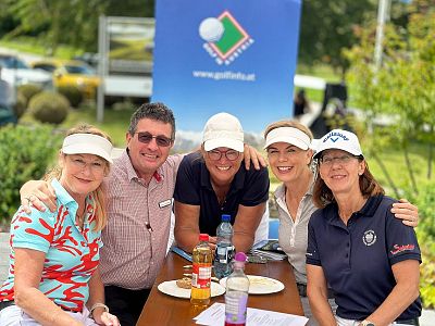 Five people sitting at a table outdoors, smiling, with golf shirts and visors. A golf-themed banner is in the background, suggesting a friendly event.