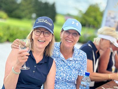 Two women enjoying a drink outdoors, wearing sports caps and casual clothes. They are smiling and seated at a table, with greenery in the background.