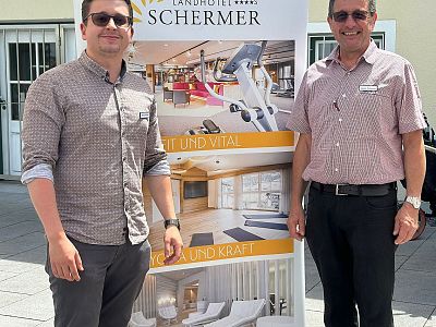 Two people stand in front of a banner advertising hotel amenities, including a fitness area and luxury rooms, outdoors on a sunny day.