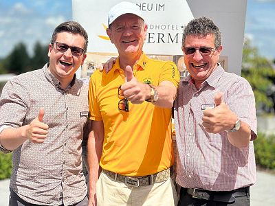 Three smiling men in casual clothing stand close together outdoors, all giving a thumbs up gesture. They appear to be enjoying a sunny day.