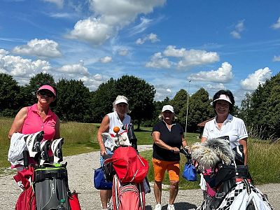 Four people in golf attire pose with their golf bags on a sunny day, surrounded by green grass and a blue sky with clouds.
