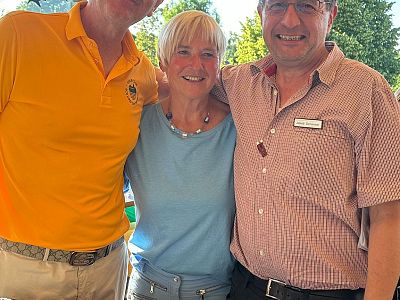 Three people smiling, one in a yellow shirt, another in blue, and the third in checkered, standing closely together outdoors on a sunny day.