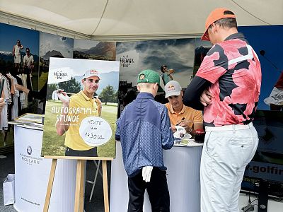 A golfer signs autographs at a booth with a poster behind, while several people stand around. It seems to be a sunny day at a golf event.