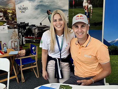 Ein lächelndes Paar in Freizeitkleidung posiert für ein Foto in einer Messeumgebung mit Golfdekorationen im Hintergrund.