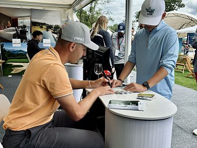 A man in a cap signs autographs for an attendee at an outdoor booth, with promotional displays and people in caps visible in the background.