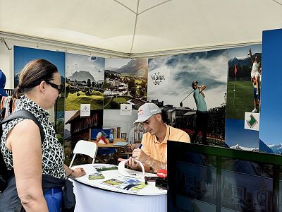 A man in an orange shirt signs an autograph at a booth while a woman looks on. The booth displays images of landscapes and golfing under a white tent.
