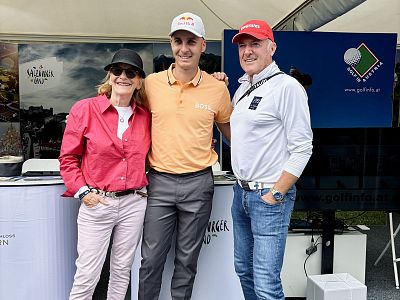 Three people smiling and posing outdoors, standing close together in casual golf attire. Behind them is a display booth with golf-related branding.
