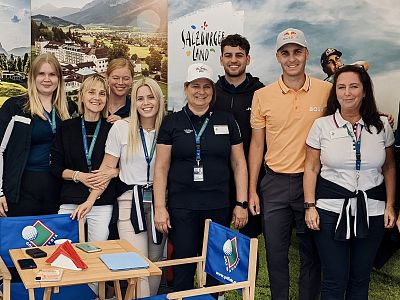 Group of eight people smiling and posing in front of promotional banners, featuring a small branded table and chairs in the foreground.
