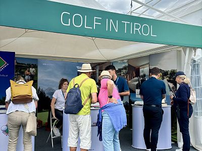 A promotional tent with a 'Golf in Tirol' sign, showing people gathered around, engaging with information materials. The setting is vibrant and interactive.