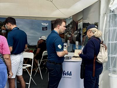 Two people conversing at a trade show booth inside a tent with brochures and displays. Other attendees are nearby, engaged with the exhibits.