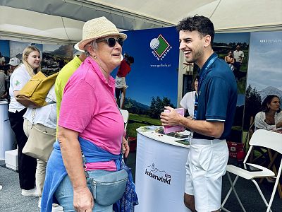 Older woman in pink shirt and hat talks to young man in navy shirt at event booth, surrounded by people and promotional materials.