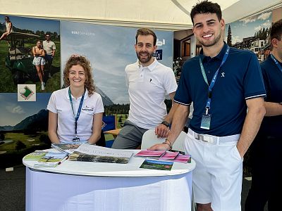 Drei Personen stehen an einem Infostand. Der Tisch ist mit Broschüren bedeckt. Sie tragen einheitliche Polo-Shirts und haben Namensschilder um den Hals.