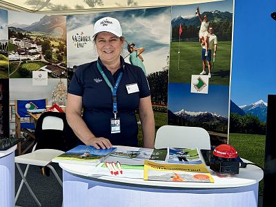 A woman at a golf-themed exhibition booth with promotional materials, wearing a cap and badge. Background features golf course and mountain images.