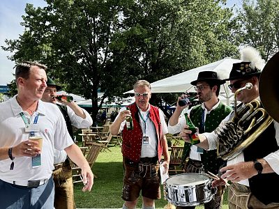 Group of people in Bavarian outfits enjoying drinks outdoors at a festival with instruments and trees in the background.