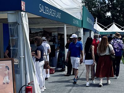 People walking and browsing booths at an outdoor exhibition under a large tent. There's a mix of casual and summer attire, indicating a sunny day.