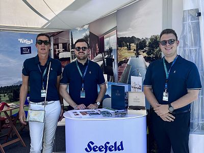 Three people stand behind a booth at an outdoor event, wearing matching navy shirts and lanyards. A sunny day with promotional banners in the background.