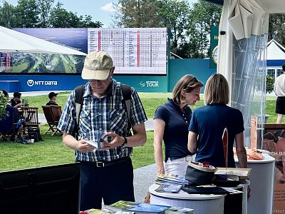 Man reads a pamphlet at a golf event booth, with others chatting nearby. A leaderboard displays scores. Expo stands and sponsor banners in the background.