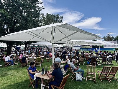 Outdoor scene with people sitting at wooden tables and chairs under large umbrellas on a grassy area, creating a lively and social atmosphere.
