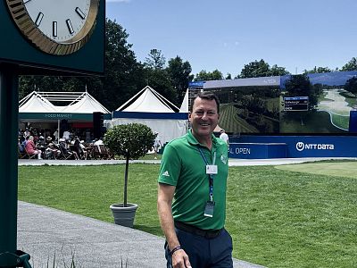 Ein Mann in einem grünen Polohemd steht lächelnd auf einem Golfplatz. Neben ihm ist eine große Uhr und im Hintergrund einige Zelte zu sehen.