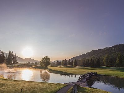 A sunrise over a tranquil golf course with a pond and a small bridge. Mist rises from the water, surrounded by trees and distant hills, creating a serene scene.