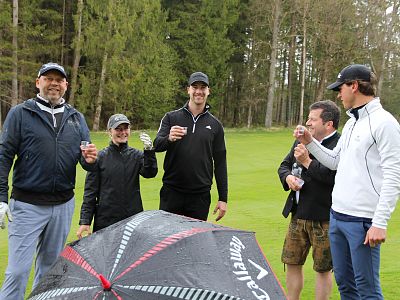 Five people stand on a golf course, toasting with drinks. They are dressed in casual sportswear, with an umbrella visible in front. Trees line the background.