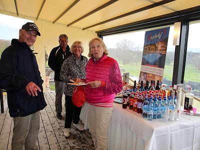 Four adults standing by a beverage table with water, soda, and wine at an outdoor event. They are dressed in outdoor clothing and seem engaged in conversation.