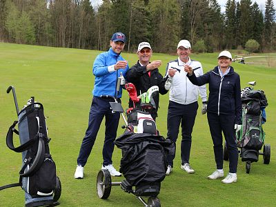 Four golfers on a course toasting with drinks, smiles on their faces, standing near their golf equipment and carts on lush green grass.