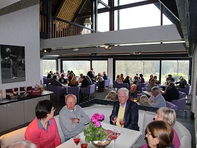 People socializing in a modern indoor space with large windows, seated at tables with drinks. Purple chairs and a relaxed atmosphere characterize the scene.
