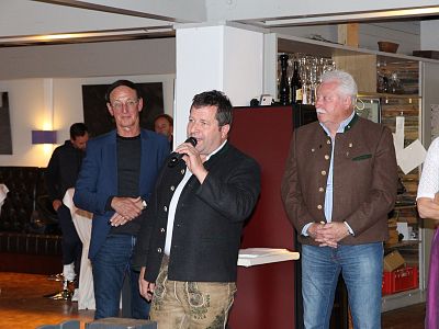 A group of people at an indoor event. A man in traditional Bavarian clothing holds a microphone. Others stand nearby, listening attentively.