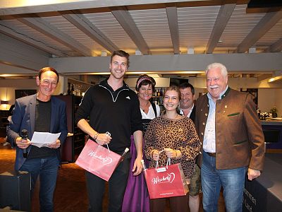 Group of six adults smiling at an indoor event, holding gift bags and wearing casual and semi-formal attire, suggesting a celebratory gathering.