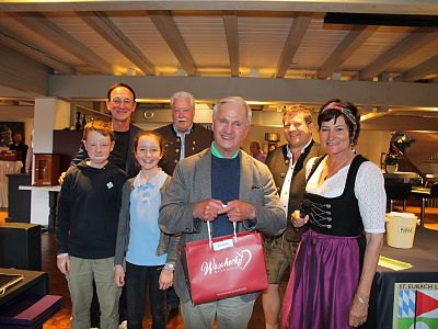 Eine Gruppe von sieben Personen steht lächelnd in einem Innenraum. Einige tragen traditionelle bayerische Trachten, während andere casual gekleidet sind. Im Hintergrund ist eine Bar zu sehen.
