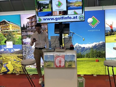 A booth promoting a golf website with banners of golf courses. A man stands checking his phone beside promotional materials and a screen with a URL.