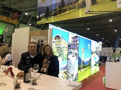 Ein Mann und eine Frau sitzen an einem Tisch auf einer Messe. Im Hintergrund sind leuchtende Werbedisplays mit Landschaftsbildern zu sehen.