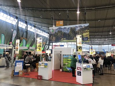 A busy trade fair booth with banners, displays, and people walking in a large hall. A backdrop shows a mountain scene under bright exhibition lights.