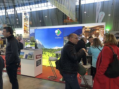 Exhibition booth with a mountain-themed backdrop, attendees interacting, promotional materials on display, and seating arrangements inside a spacious hall.