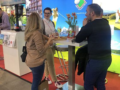 Three individuals converse at a trade show booth with colorful posters and a snack bowl, reflecting an engaging and vibrant environment.