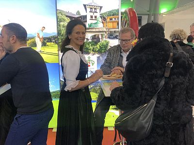 People at a fair booth, one in traditional attire, interacting with visitors against a backdrop showcasing scenic landscapes.