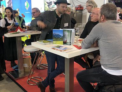 Several people at a trade show booth discussing over a high table with promotional materials around them, some seated and some standing, in a busy exhibition setting.