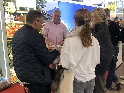 Four people are conversing at an exhibition booth, with a display screen in the background. They are leaning over a table, possibly discussing materials.