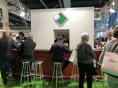 People gather at a white exhibition booth with a green and red logo. Some are seated on stools, others stand, engaging in conversations.
