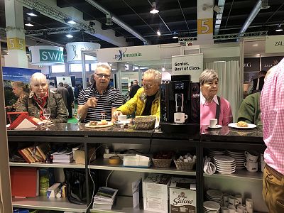 Several people stand at a counter with coffee machines, enjoying drinks and snacks at an event with booths and displays in the background.