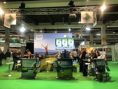 Crowded expo booth with golf theme, featuring banners, screens, and attendees browsing displays on a green carpet.
