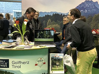 People gather at a booth labeled 'Golfherz Tirol' with brochures, a laptop, and tulips, against a mountain backdrop.
