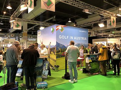 People gather around a booth at a trade fair. The booth promotes golfing in Austria, displaying scenic golf course images. Attendees engage and explore materials.