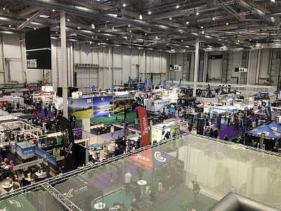 A bustling exhibition hall with numerous booths and banners, filled with people walking around and engaging with exhibitors under bright lights.