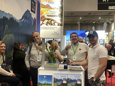 Four people at a tourism booth, smiling and holding drinks, with brochures on the table. The booth displays mountain scenery posters in the background.